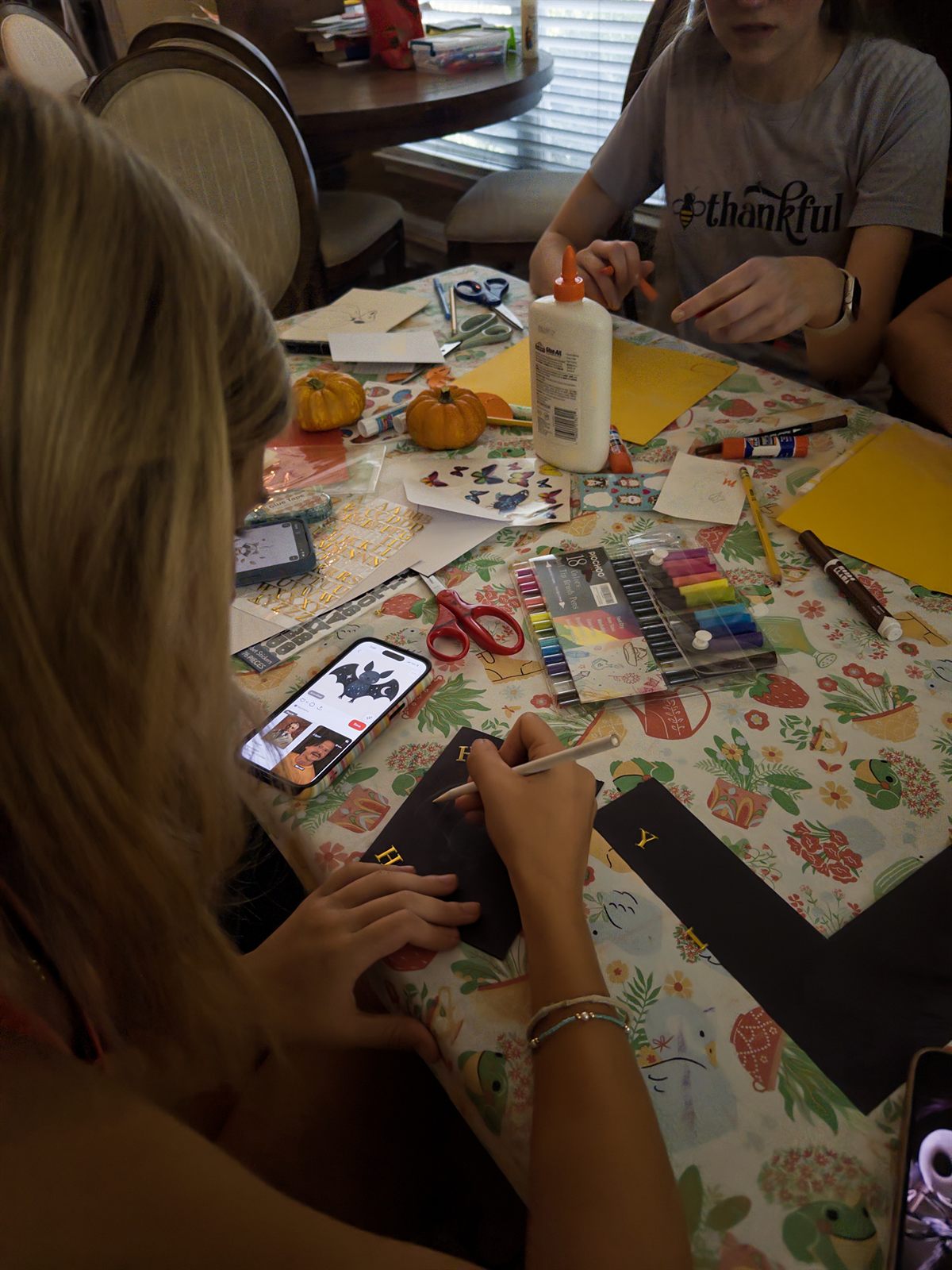 A member lettering a black card in gold, with a drawing pulled up on her phone for reference